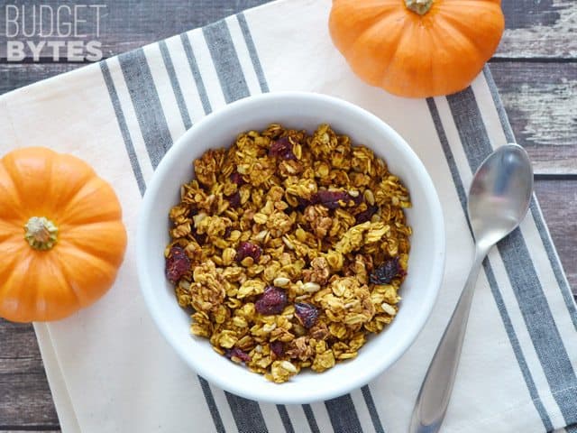 Top view of a bowl of Pumpkin Spice Granola, spoon, napkin and small pumpkins on the side