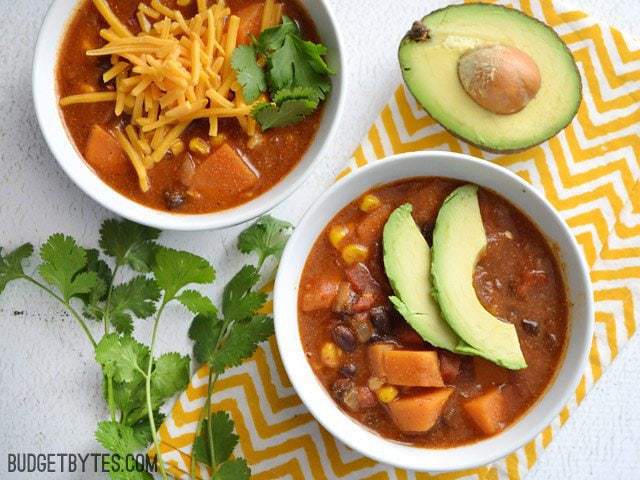 Top view of two bowls of Sweet Potato Tortilla Soup, one garnished with avocado and one with cheese and cilantro. Yellow chevron napkin, half an avocado and cilantro on the side for staging.