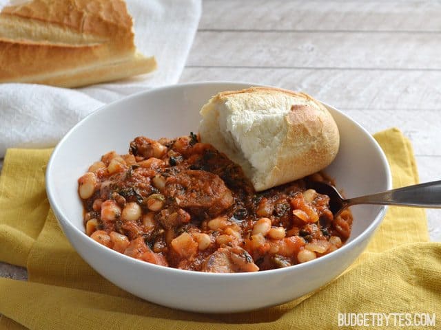 Side view of a bowl of White Beans with Tomato and Sausage with a slice of bread, sitting on a yellow napkin