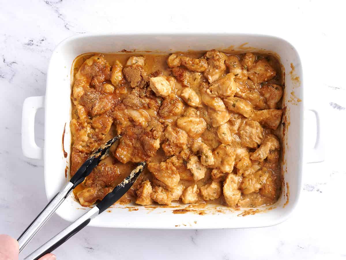Tongs tossing Thai peanut chicken breast in a baking dish.