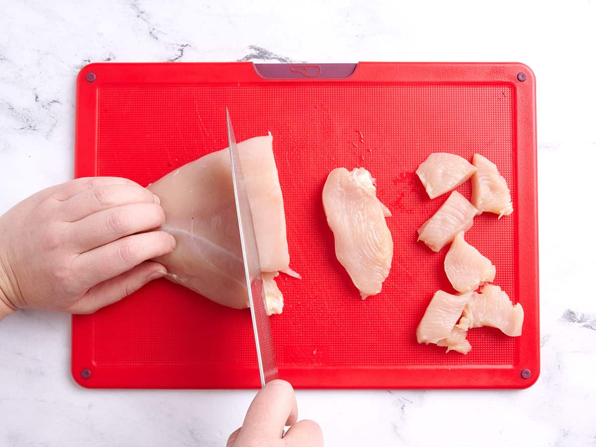 A knife slicing a chicken breast into pieces on a cutting board.