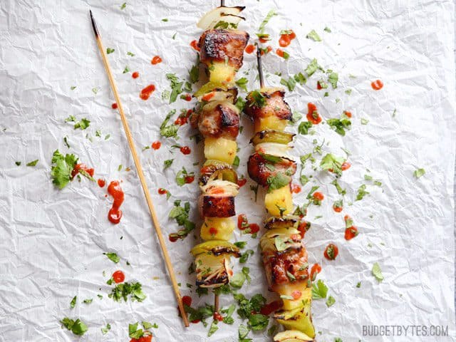 These incredibly simple Pineapple Pork Kebabs have flame licked flavor but don't require an outdoor grill. Fresh, bright, and perfect for summer! Budgetbytes.com Two Pineapple Pork Kebabs garnished with cilantro and sriracha next to an empty skewer