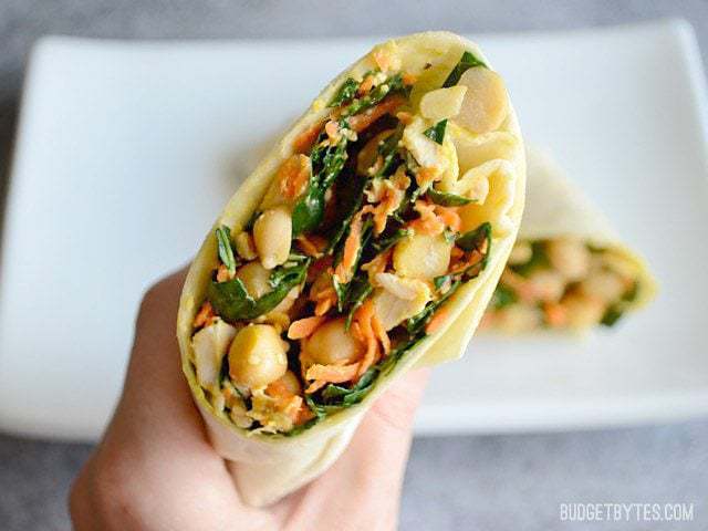 This easy Kale Chicken Caesar Wrap is a great no-cook, no-reheat lunch idea. A big deli-style wrap made at home for a LOT less money. Budetbytes.com A hand holding half of a kale chicken caesar wrap close to the camera