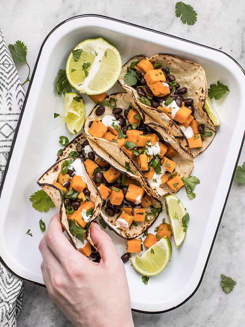 These light and refreshing Sweet Potato Tacos are filled with a simple sweet potato and black bean hash, and topped with a tangy lime crema. Budetbytes.com A hand picking up a sweet potato taco out of the serving dish