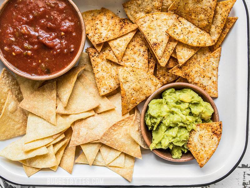 These Homemade Baked Tortilla Chips are fast, easy, super crunchy, a great way to use up leftover tortillas, and a great alternative to store bought chips. Budgetbytes.com Close up of Homemade Baked Tortilla Chips with bowls of salsa and guacamole