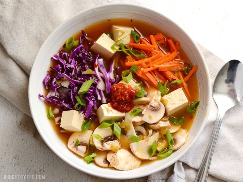 This easy hot and sour soup is light on the stomach, but big on flavor! The spicy and tangy broth is full of colorful vegetables and tender cubes of tofu. BudgetBytes.com Overhead view of a bowl full of hot and sour soup with vegetables and cubed tofu, chili garlic sauce in the center of the bowl