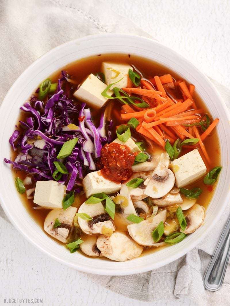 This easy hot and sour soup is light on the stomach, but big on flavor! The spicy and tangy broth is full of colorful vegetables and tender cubes of tofu. BudgetBytes.com Overhead view of a bowl full of hot and sour soup with vegetables and tofu. Garnished with a dab of chili garlic sauce.