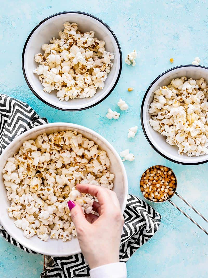 How to make perfect fluffy and crunchy stovetop popcorn with just a couple of ingredients and one pan. The perfect, fast, and inexpensive snack! BudgetBytes.com A hand picking up a handful of popcorn from a large bowl, two smaller bowls on the side