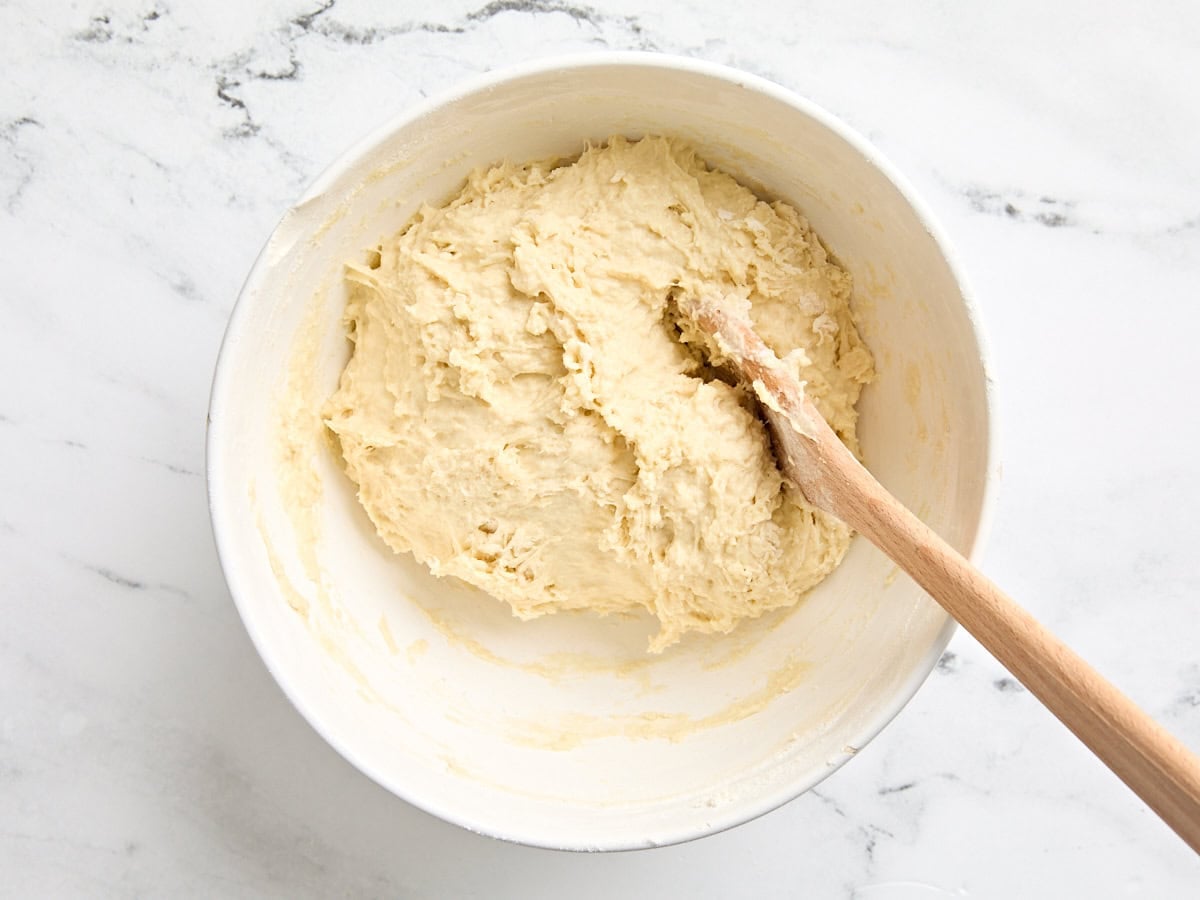 A wooden spoon mixing a ball of naan dough in a mixing bowl.