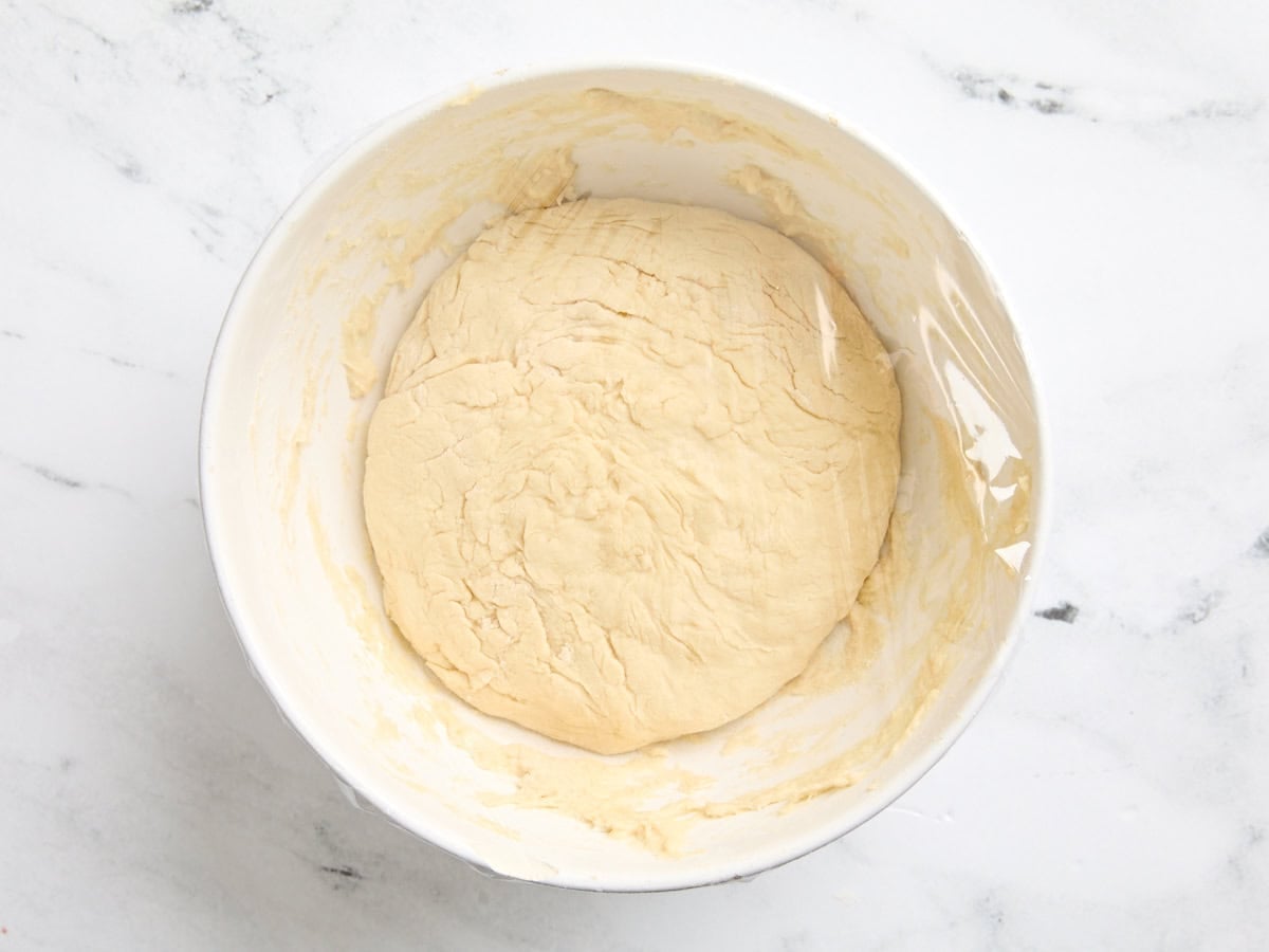 Homemade naan dough in a mixing bowl covered with plastic wrap to rise.