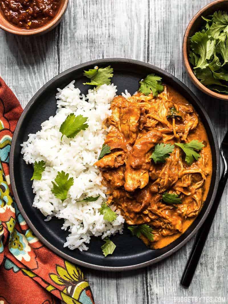 This Slow Cooker Chicken Tikka Masala boasts a rich and aromatic sauce, and tender juicy chicken. Make four servings for the price of one take out! Budgetbytes.com Slow Cooker Chicken Tikka Masala on a black plate with rice and cilantro.