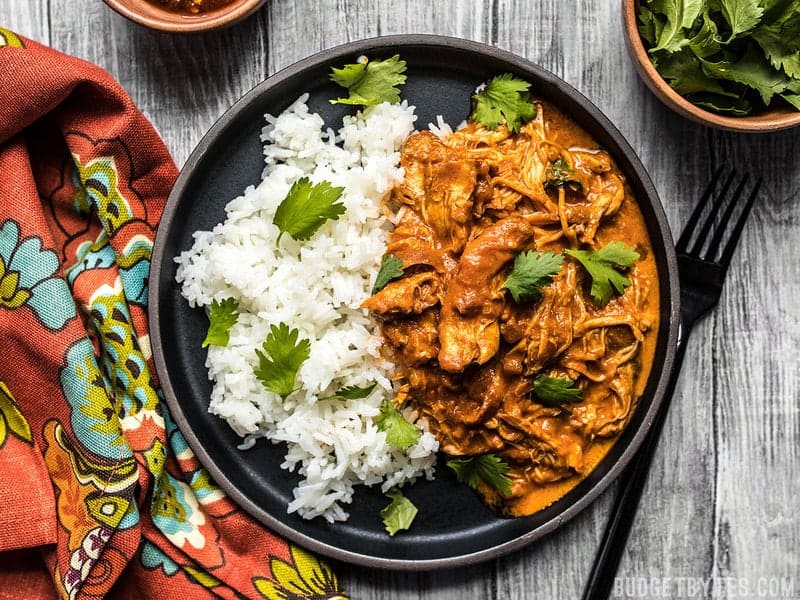 This Slow Cooker Chicken Tikka Masala boasts a rich and aromatic sauce, and tender juicy chicken. Make four servings for the price of one take out! Budgetbytes.com A plate full of slow cooker chicken tikka masala served with rice and cilantro, a colorful napkin on the side.