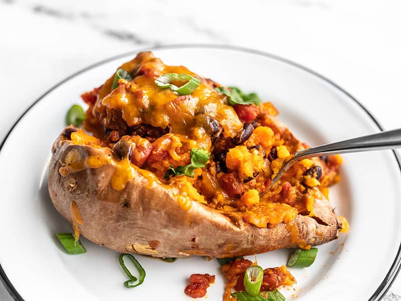 Tender baked sweet potatoes smothered with turkey and bean chili, then topped with melty cheddar. Turkey Chili Smothered Sweet Potatoes are an easy dinner! BudgetBytes.com A fork digging into a turkey chili smothered sweet potato, on a white plate