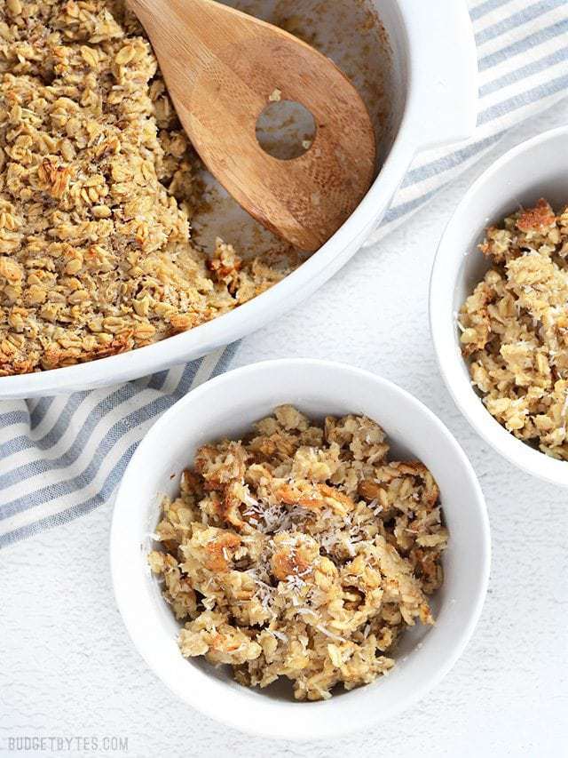 Rich and slightly sweet, this Banana Coconut Baked Oatmeal tastes indulgent, but is a balanced, fiber filled breakfast that will keep you full all morning! Budetbytes.com Banana Coconut Baked Oatmeal served into two bowls, casserole dish with a wooden spoon on the side