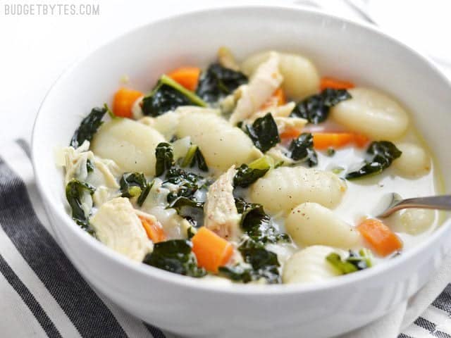 Take your classic chicken soup up a notch with the addition of kale, gnocchi, and cream. Creamy Chicken Gnocchi Soup with Kale is a warm and comforting meal! Budetbytes.com Side view of a bowl of creamy chicken gnocchi soup with kale, a spoon in the center