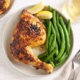 Overhead view of a baked chicken leg on a plate with green beans and lemon wedges.