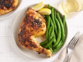 Overhead view of a baked chicken leg on a plate with green beans and lemon wedges.