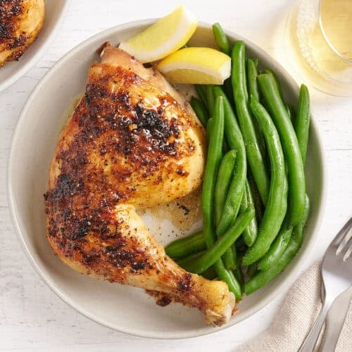 Overhead view of a baked chicken leg on a plate with green beans and lemon wedges.