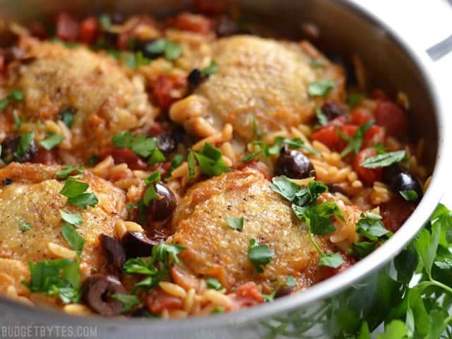 This Chicken with Orzo and Olives simmers together in one skillet for maximum flavor and minimum cleanup. A one-pot easy weeknight dinner! Budetbytes.com Close up side view of Chicken with Orzo and Olives in the skillet, garnished with parsley