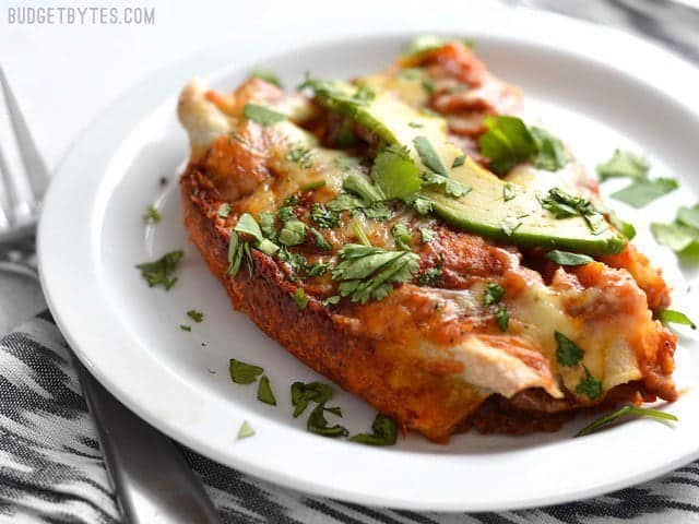 These easy Bean and Cheese Enchiladas are anything but boring. A few extra details take them from a simple to spectacular weeknight dinner. BudgetBytes.com Two bean and cheese enchiladas on a white plate, sitting on a black and white cloth napkin.