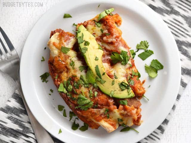 These easy Bean and Cheese Enchiladas are anything but boring. A few extra details take them from a simple to spectacular weeknight dinner. BudgetBytes.com Two baked enchiladas on a plate, topped with an avocado slice and chopped cilantro.