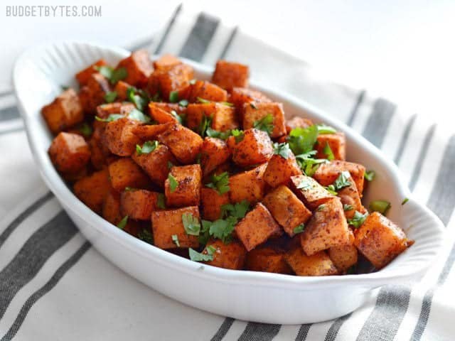 Chili Roasted Sweet Potatoes are an easy and flavorful side dish for any southwest inspired meal. Perfect for busy weeknights and meal prep! BudgetBytes.com Side view of a white serving dish full of Chili Roasted Sweet Potatoes garnished with cilantro