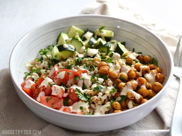 This Mediterranean Farro Salad with Spiced Chickpeas is a flavorful and filling vegan meal packed with tons of fiber and nutrients. Perfect for meal prep! Budetbytes.com Front view of a bowl of Mediterranean Farro Salad topped with tahini dressing and parsley