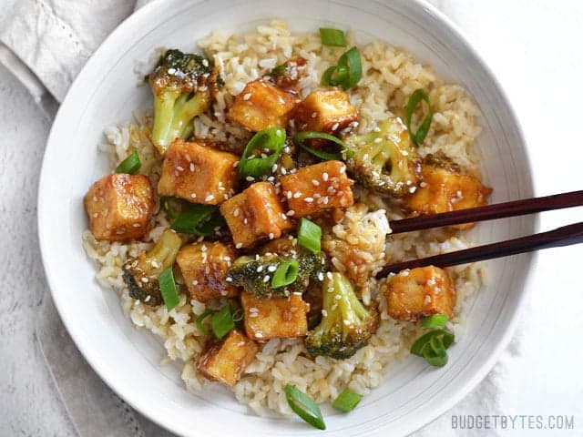 This Pan Fried Sesame Tofu is seriously crispy and drenched in a tangy sesame sauce. Broccoli florets and cooked rice make it a meal. Step by step photos. Budgetbytes.com Chopsticks digging into a bowl of Pan Fried Sesame Tofu with Broccoli and brown rice.