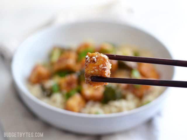 This Pan Fried Sesame Tofu is seriously crispy and drenched in a tangy sesame sauce. Broccoli florets and cooked rice make it a meal. Step by step photos. Budgetbytes.com Close up of one cube of pan fried sesame tofu with sesame seeds on top.