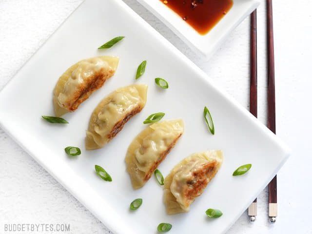 Filling, folding, frying, and steaming your own Pork Gyoza at home is not only fun, but it costs just pennies on the dollar compared to restaurants. BudgetBytes.com Four Pork Gyoza on a plate, sprinkled with green onion, next to a dish of soy sauce.