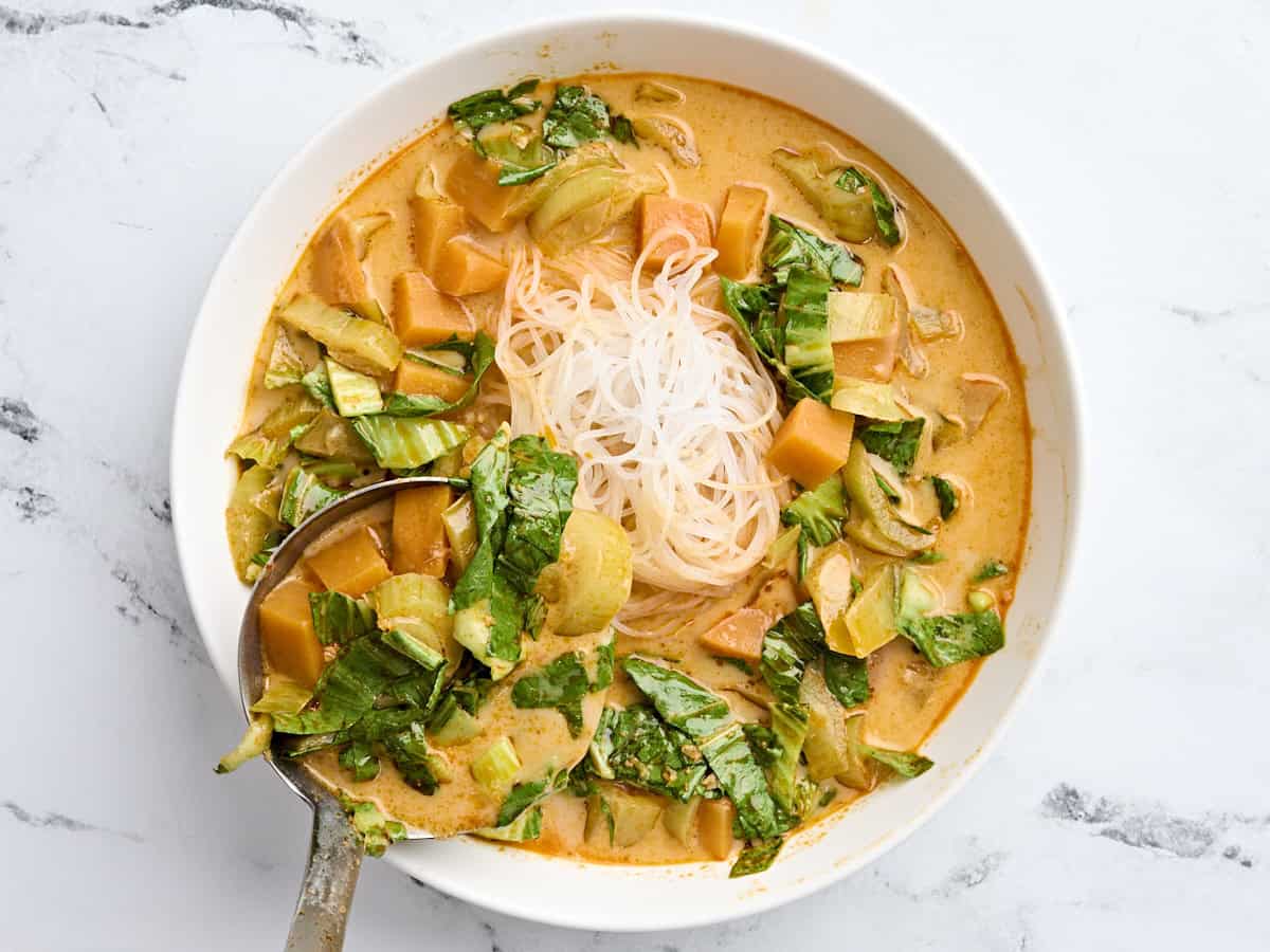 Thai curry vegetable soup being spooned over cooked noodles in a bowl.