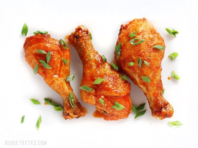 Sweet and spicy honey sriracha chicken drumsticks baked to perfection in the oven! Change up the sauce for a different meal every week! BudgetBytes.com Three Crispy Baked Honey Sriracha Chicken Drumsticks on a white plate, garnished with green onion
