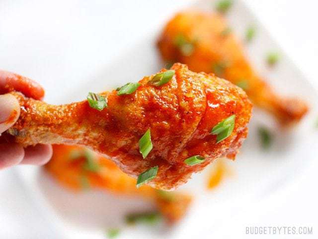 Sweet and spicy honey sriracha chicken drumsticks baked to perfection in the oven! Change up the sauce for a different meal every week! BudgetBytes.com One honey sriracha baked drumstick held close to the camera, garnished with green onions