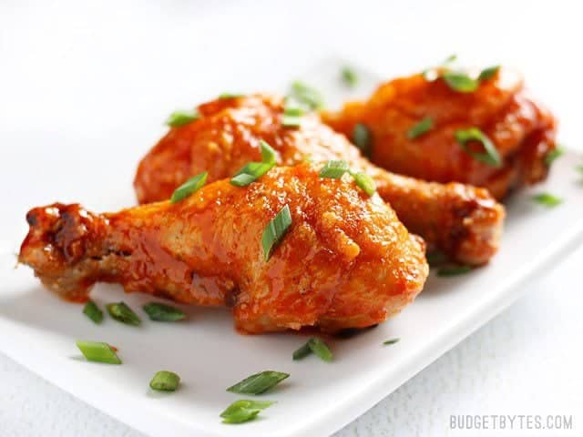 Sweet and spicy honey sriracha chicken drumsticks baked to perfection in the oven! Change up the sauce for a different meal every week! BudgetBytes.com Side view of Crispy Baked Honey Sriracha Chicken Drumsticks on a white rectangular plate