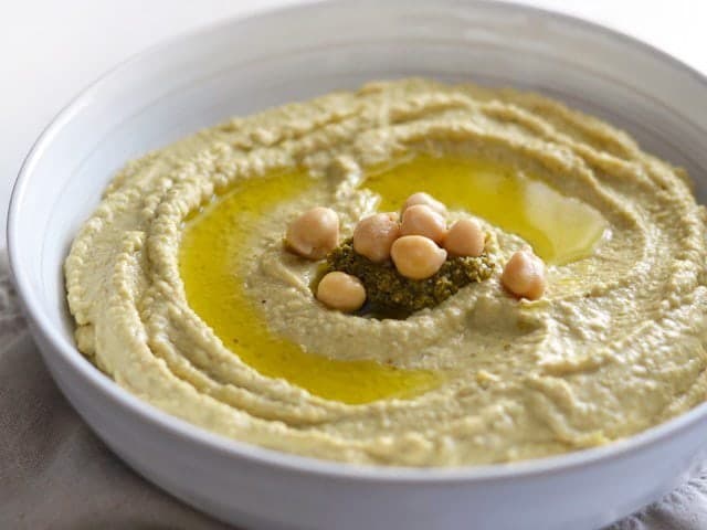 Spiking a basic hummus recipe with a dollop of basil hummus adds big flavor. Use this herby pesto hummus for sandwiches, dipping, on pasta, and more! BudgetBytes.com Close up side view of a bowl of Pesto Hummus garnished with oil, pesto, and chickpeas