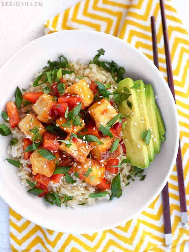 Sweet Chili Tofu Bowls are an easy, fresh, and flavorful vegan weeknight dinner, packed with vibrant colors and flavors. Perfect for meal prep! BudgetBytes.com Overhead view of a Sweet Chili Tofu Bowl on a yellow zig zag napkin, chopsticks on the side
