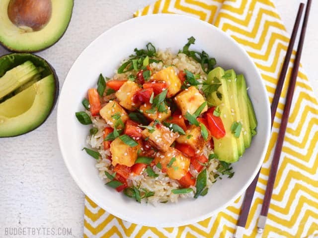 Sweet Chili Tofu Bowls are an easy, fresh, and flavorful vegan weeknight dinner, packed with vibrant colors and flavors. Perfect for meal prep! BudgetBytes.com Overhead view of a Sweet Chili Tofu Bowl on a yellow and white napkin, a cut open avocado on the side