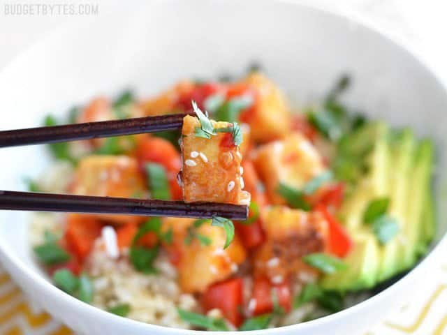 Sweet Chili Tofu Bowls are an easy, fresh, and flavorful vegan weeknight dinner, packed with vibrant colors and flavors. Perfect for meal prep! BudgetBytes.com Close up of chopsticks holding one tofu cube coated in sweet chili sauce, the bowl in the background