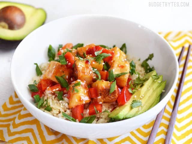 Sweet Chili Tofu Bowls are an easy, fresh, and flavorful vegan weeknight dinner, packed with vibrant colors and flavors. Perfect for meal prep! BudgetBytes.com Front view of Sweet Chili Stir Fried Tofu Bowl on a napkin, chopsticks on the side, avocado in the back