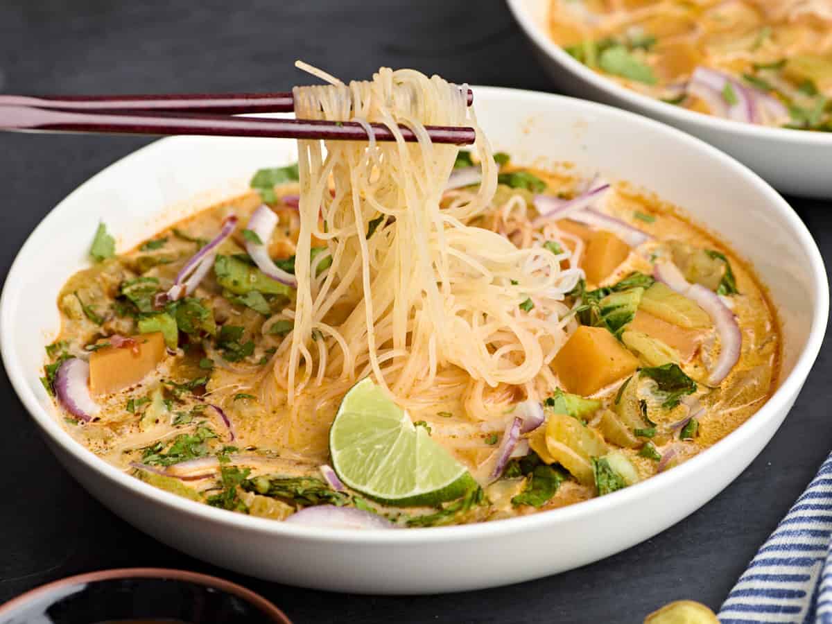 Side view of a homemade thai red curry soup in a bowl, with chopsticks taking some noodles from the bowl.