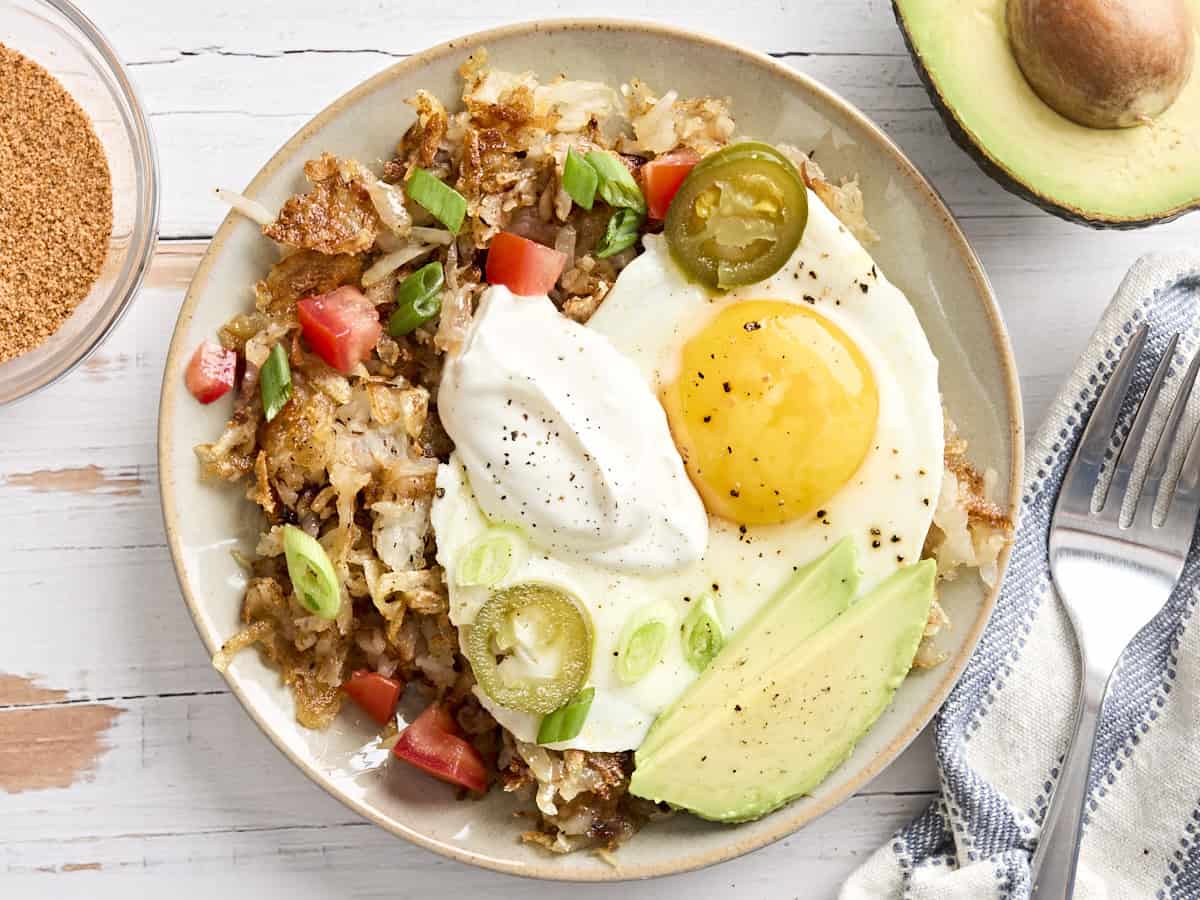 Overhead view of homemade hash browns on a plate, topped with a fried egg, sour cream, avocado, diced tomato, and sliced jalapeño,