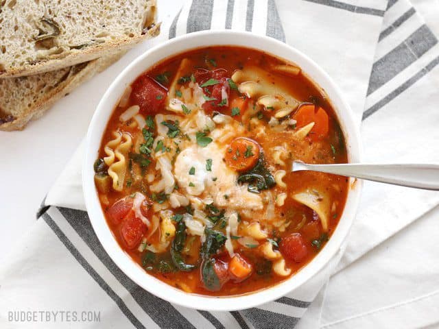 This Garden Vegetable Lasagna Soup is packed full of vegetables and lasagna flavor, plus a hidden dollop of melty three cheese ricotta blend in the middle! BudgetBytes.com Overhead view of a bowl full of Garden Vegetable Lasagna soup with a spoon in the center, bread on the side
