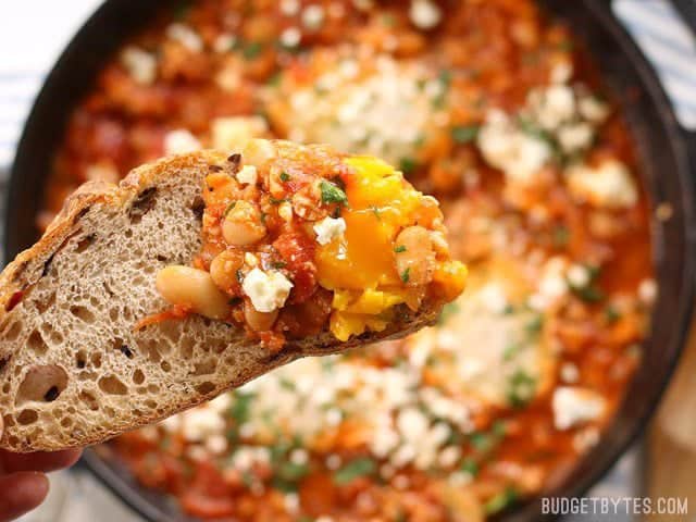 Eggs poached in a rich and smoky tomato sauce speckled with white beans, this Smoky White Bean Shakshuka is a filling and inexpensive breakfast or dinner! BudgetBytes.com Close up of a slice of bread with some Smoky White Bean Shakshuka on the end.