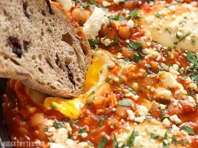 Eggs poached in a rich and smoky tomato sauce speckled with white beans, this Smoky White Bean Shakshuka is a filling and inexpensive breakfast or dinner! BudgetBytes.com Close up of a slice of bread being dipped into a runny. yolk in the Smoky White Bean Shakshuka