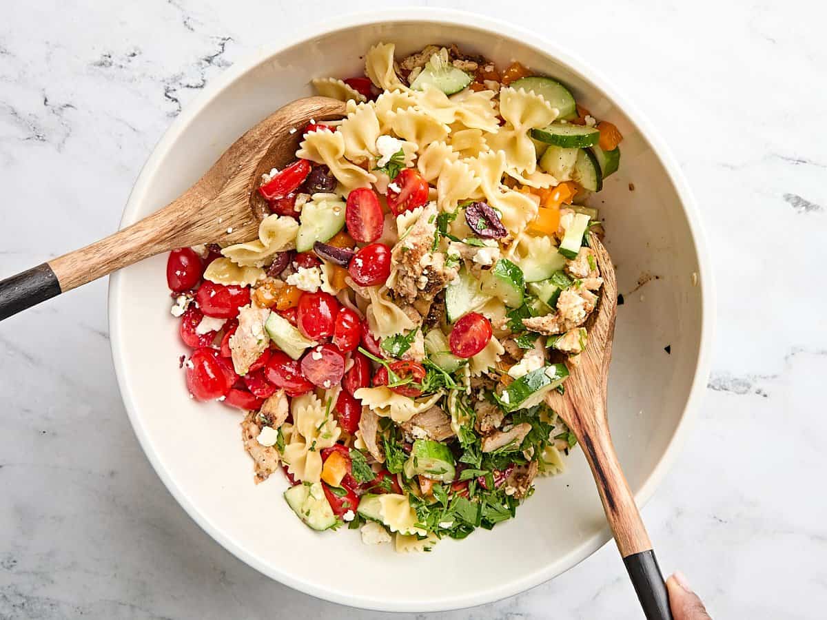 Greek Chicken Pasta Salad is the perfectly refreshing and filling summer meal with its medley of vegetables and tangy lemon garlic dressing. BudgetBytes.com Two wooden spoons tossing a greek chicken salad in a bowl.