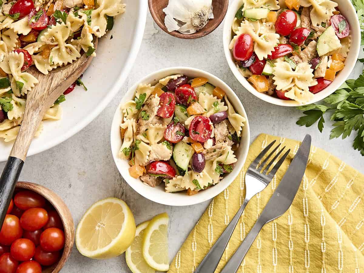 Overhead view of a Greek chicken pasta salad recipe in two bowls next to a larger platter.