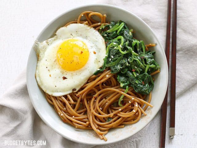 These Easy Sesame Noodles with Wilted Greens are a simple vegetarian meal with big flavor and plenty of options for customization. BudgetBytes.com Overhead view of a bowl full of Sesame Noodles with Wilted Greens topped with a fried egg and chopsticks on the side