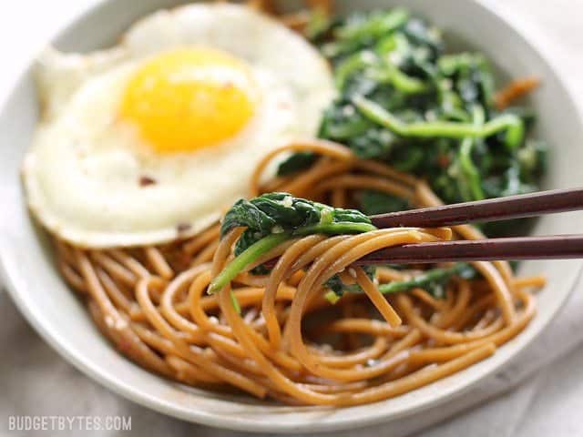 These Easy Sesame Noodles with Wilted Greens are a simple vegetarian meal with big flavor and plenty of options for customization. BudgetBytes.com Close up side view of a bowl of Sesame Noodles with Wilted Greens with chopsticks picking up some noodles