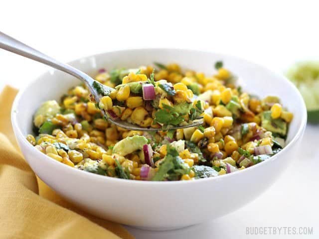 Warm Corn and Avocado Salad makes the perfect light and fresh side dish for enchiladas, grilled meats, tacos, or any summer meal. BudgetBytes.com Front view of a bowl full of Warm Corn and Avocado Salad with a fork lifting a bite