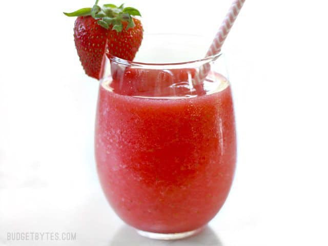 This Strawberry Rosé Slush is a fast and inexpensive frozen drink made with wine and fruit that you can enjoy all summer. No fancy ingredients required! BudgetBytes.com Side view of a Strawberry Rosé Slush with a pink paper straw and fresh strawberry on the rim.